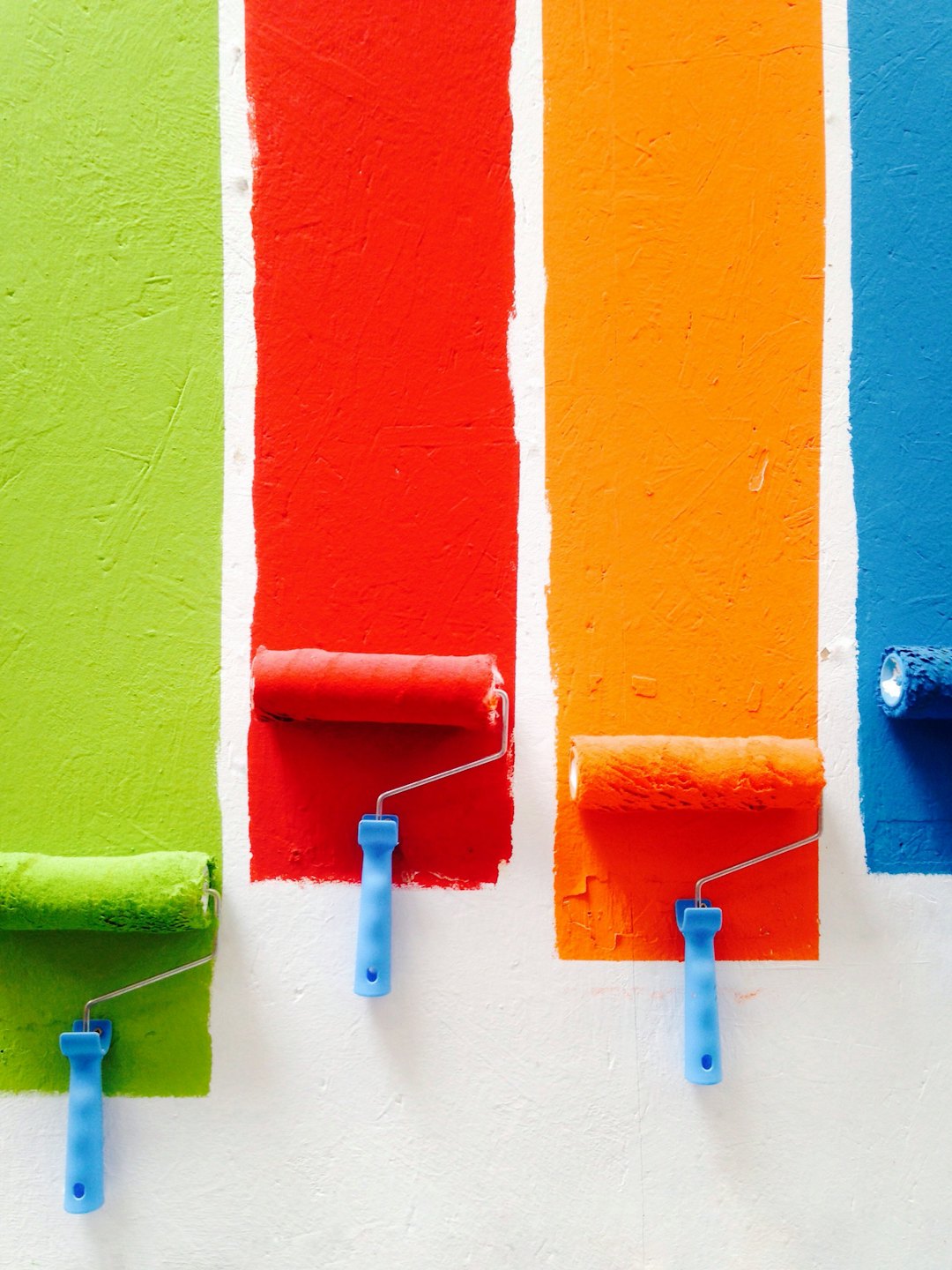 Four paint rollers rest against a white wall below fresh paint stripes.